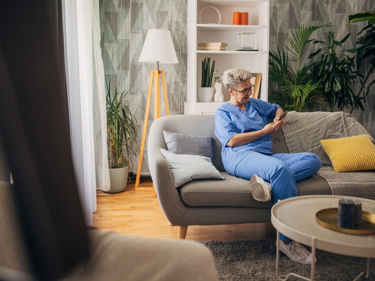 Person in scrubs sitting on a sofa at home using a smartphone in a relaxed living room